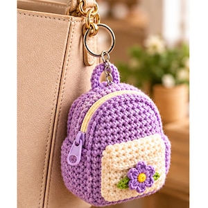 May include: A small, purple, crocheted backpack keychain featuring a cream-colored front pocket adorned with a purple flower with a yellow center. The backpack has a light purple zipper pull and is attached to a gold-toned metal ring and chain.