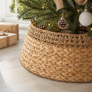 May include: A round, tapered Christmas tree collar constructed from woven natural seagrass. The base features a dense, braided pattern, while the upper section displays a decorative, open-weave lattice design in a matching light tan color.