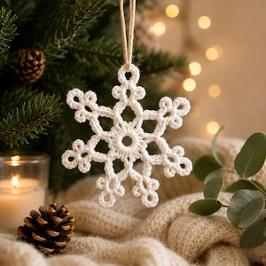 Op de afbeelding: Een wit gehaakt sneeuwvlokornament met ingewikkelde kantachtige patronen en een centrale ronde opening. De sneeuwvlok hangt aan een dunne, lichtbruine jute lus en toont gedetailleerd stikwerk en een symmetrisch ontwerp met zes punten.
