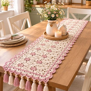 Puede incluir: Un camino de mesa rectangular de ganchillo con un intrincado patrón floral en crema y rosa con un borde festoneado. El camino está rematado con borlas de hilo alternas en color crema y rosa a lo largo de los extremos cortos.
