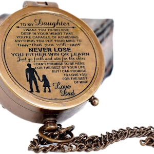 May include: A round, antique-style brass compass with a hinged lid featuring an engraved message to a daughter, a silhouette of a parent and child, and a metal chain. The text reads: TO MY DAUGHTER, I WANT YOU TO BELIEVE DEEP IN YOUR HEART THAT YOU'RE CAPABLE OF ACHIEVING ANYTHING YOU PUT YOUR MIND TO, THAT YOU WILL NEVER LOSE, YOU EITHER WIN OR LEARN, JUST GO FORTH AND AIM FOR THE SKIES, I CAN'T PROMISE TO BE HERE FOR THE REST OF YOUR LIFE, BUT I CAN PROMISE TO LOVE YOU FOR THE BEST OF MINE, LOVE DAD.