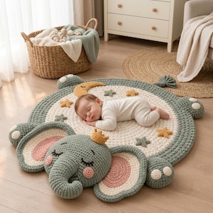 May include: A round, crocheted play mat featuring a large, sage green elephant head with pink inner ears and a small yellow crown. The center is cream-colored with scattered yellow and green stars and a crescent moon, providing a soft surface.