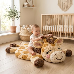 May include: A large, flat, crocheted giraffe floor mat featuring a cream-colored body with tan spots, dark brown hooves, and a plush, smiling giraffe head with closed eyes, pink cheeks, and a small tuft of brown yarn hair on the top of its head.