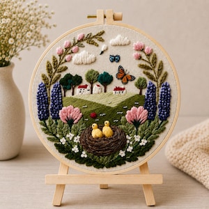 May include: A circular embroidery hoop art piece featuring a landscape scene with two yellow chicks in a brown nest, green hills, white houses, purple lupine flowers, pink blossoms, a monarch butterfly, and white clouds on a beige fabric background.