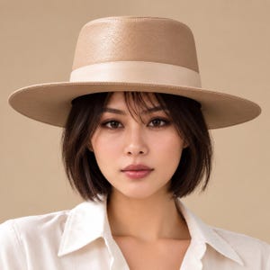 May include: A structured fedora hat in a light tan colour with a textured finish. The hat features a wide, flat brim and a smooth, tonal band encircling the base of the crown. The material appears firm, maintaining a clean, symmetrical shape.