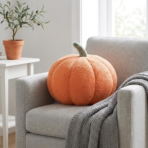 May include: A round, orange pumpkin-shaped cushion with a textured, fuzzy surface and a short, green stem at the top. The cushion features deep, vertical ridges that mimic the natural segments of a pumpkin, creating a soft, plush appearance.