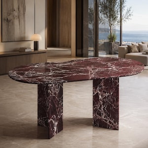 May include: An oval dining table crafted from deep burgundy marble with prominent white veining. The table features a smooth, polished surface supported by two thick, rectangular slab legs made of the same matching dark red marble material.