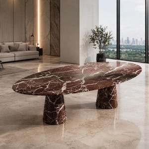 Può includere: Un tavolo da pranzo ovale realizzato in marmo rosso intenso con venature bianche prominenti. Il tavolo presenta una superficie liscia e lucida, sostenuta da due spessi piedistalli conici che riprendono il motivo del marmo del piano.
