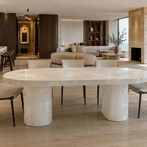 Puede incluir: Una mesa de comedor ovalada con una superficie de piedra lisa en color blanco roto y dos bases de pedestal cilíndricas y gruesas. La mesa está rodeada por cuatro sillas con asientos tapizados en beige claro y marcos de color marrón oscuro.