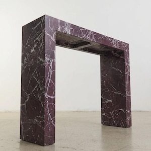 Pode incluir: Uma mesa consola retangular com acabamento em mármore roxo profundo e veios brancos proeminentes. A estrutura consiste em dois painéis laterais verticais que suportam uma peça superior horizontal, criando um design geométrico aberto.