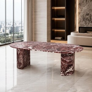 Puede incluir: Una mesa de comedor ovalada con una superficie de mármol rojo intenso con vetas blancas. La mesa se apoya sobre dos bases de pedestal gruesas y curvas de mármol rojo a juego que proporcionan una base sólida para la superficie de piedra pulida.