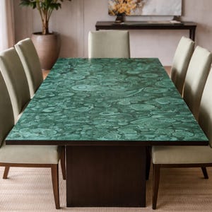 Puede incluir: Una mesa de comedor rectangular con una superficie de patrón de malaquita verde vibrante, bordes de color marrón oscuro y una base de pedestal sólida, rodeada por ocho sillas tapizadas en tela beige claro con patas de madera oscura.