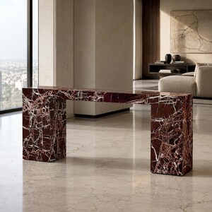 May include: A rectangular console table constructed from deep red marble with prominent white veining. The piece features a flat top surface supported by two thick, block-style vertical legs, creating a continuous, solid, and geometric structure.