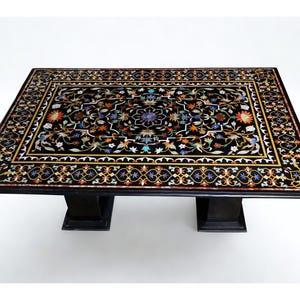Puede incluir: Una mesa rectangular de piedra negra con un intrincado diseño de incrustaciones florales y detalles en piedras de colores. El tablero muestra un medallón central rodeado por un patrón de borde detallado, sobre dos bases de pedestal negras.