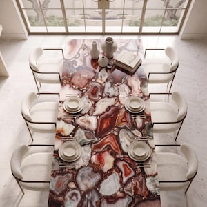 May include: A rectangular dining table with a polished surface made of multicoloured agate slices in shades of red, brown, and white. The table is surrounded by six cream-coloured upholstered chairs with thin metal frames and set with white plates.