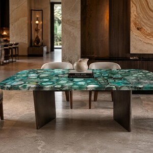 Puede incluir: Una mesa de comedor rectangular con una superficie pulida hecha de rodajas de ágata verde con variados patrones circulares. La mesa está sostenida por dos bases de pedestal oscuras y sólidas, rodeada por cuatro sillas tapizadas con patas de madera.