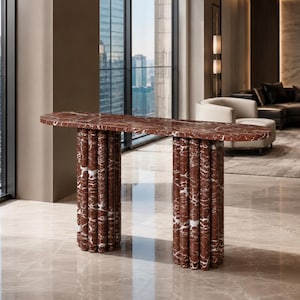 May include: A console table crafted from deep red marble with prominent white veining. The piece features a long, thin, oval-shaped top supported by two substantial bases, each composed of a cluster of vertical, cylindrical marble columns.