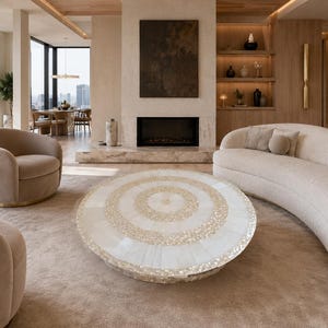 Op de afbeelding: Een ronde salontafel met een oppervlak ingelegd met witte en crèmekleurige parelmoertegels in concentrische cirkelpatronen, rustend op een lage voet, geplaatst in een woonkamer met beige meubels.