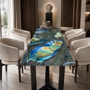 Puede incluir: Una mesa de comedor rectangular con un tablero de piedra pulida con un patrón de mármol azul y dorado, apoyada sobre una base de metal negro con marco angular, rodeada por cuatro sillas tapizadas de color crema con respaldos curvos.