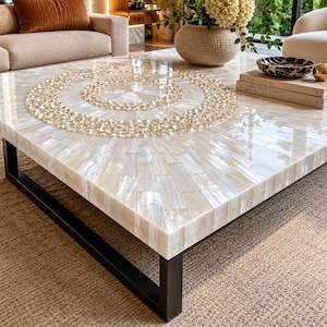 Pode incluir: Uma mesa de centro quadrada com uma superfície de azulejos de madrepérola branca e um padrão circular central incrustado. A mesa é sustentada por uma estrutura de metal preto. A superfície é brilhante e reflete a luz, destacando a textura iridescente.