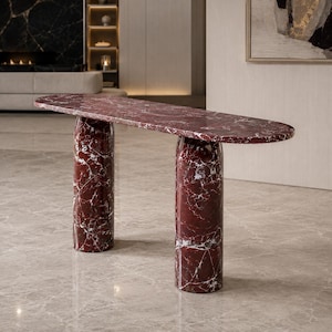 Può includere: Un tavolo console realizzato in marmo rosso intenso con venature bianche prominenti. Il pezzo presenta un piano ovale lungo e sottile, sostenuto da due spesse colonne cilindriche che condividono la stessa finitura in pietra lucida e motivo.