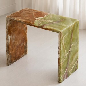 May include: A waterfall-style console table crafted from polished onyx stone. The piece features a smooth, rectangular surface and two vertical side panels, showcasing a natural color gradient from deep amber-brown to light sage green.