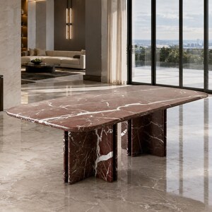 May include: A large dining table featuring a polished deep red marble top with prominent white veining. The table is supported by a substantial base constructed from matching red marble panels, creating a sturdy and cohesive structural design.