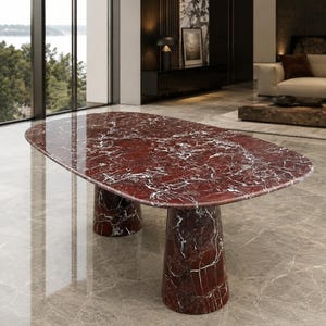 May include: An oval dining table crafted from deep red marble with prominent white veining. The table features a smooth, polished surface and is supported by two thick, cylindrical pedestal bases made from the same matching red marble material.