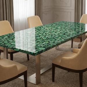 Puede incluir: Una mesa de comedor rectangular con una superficie de piedra de malaquita verde pulida con un patrón circular, sostenida por una base de metal plateado, rodeada por seis sillas tapizadas en beige con patas de madera oscura.