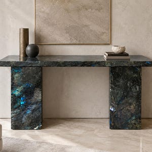 May include: A rectangular console table crafted from dark, polished stone featuring iridescent blue and black mineral inclusions. The table is supported by two thick, matching stone slab legs and holds a tall bronze cylinder, a black sphere, and a small bowl.