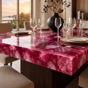 Puede incluir: Una mesa de comedor con una superficie de piedra ágata magenta pulida con un patrón cristalino natural. La mesa está dispuesta con copas de cristal transparente, platos de cerámica blanca y tenedores de metal dorado, con un gran jarrón negro.