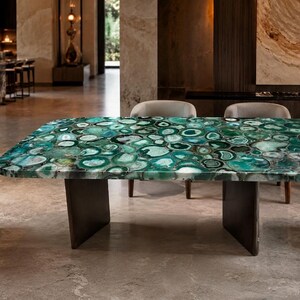 Puede incluir: Una mesa de comedor rectangular con una superficie pulida hecha de numerosas rodajas de ágata verde y azul verdoso. El tablero está sostenido por dos bases de pedestal rectangulares, oscuras y sólidas. La mesa está rodeada por cuatro sillas.