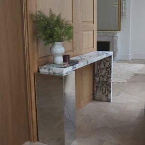 May include: A console table featuring a white and black marble top and side panel, paired with a reflective silver-toned metal base. A white textured ceramic vase containing green foliage sits on the surface alongside a small dark book.