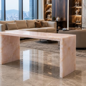 May include: A rectangular console table crafted from polished light pink stone with a smooth, reflective surface. The piece features a waterfall design with two vertical side panels supporting a flat top, showcasing natural veining throughout.