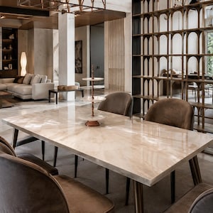 May include: A rectangular dining table with a polished light beige stone top and tapered legs, surrounded by six upholstered brown chairs. A three-tiered metal serving stand sits in the centre of the table, which is set in a large open room.