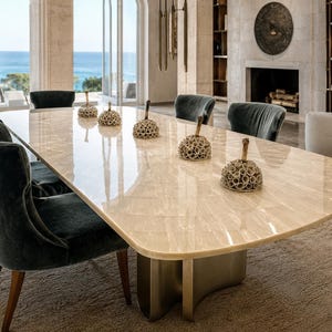 Puede incluir: Una mesa de comedor grande de forma ovalada con una superficie de piedra pulida de color beige claro y una base metálica curva. Cuatro objetos decorativos texturizados en forma de cúpula con tallos están alineados en el centro.