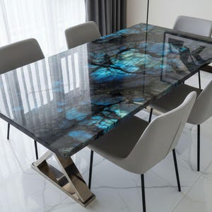 Puede incluir: Una mesa de comedor rectangular con un tablero de piedra labradorita pulida con patrones cristalinos en azul profundo y negro. La mesa se apoya sobre una base de metal plateado angular y reflectante, rodeada por seis sillas tapizadas en gris claro.