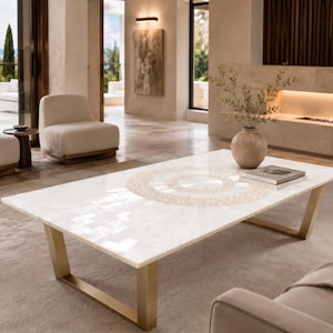 Puede incluir: Una mesa de centro rectangular con una superficie de azulejos blancos que presenta un patrón circular dorado en el centro. La mesa se apoya sobre dos patas angulares de metal dorado cepillado. Un jarrón de cerámica y un libro están sobre ella.
