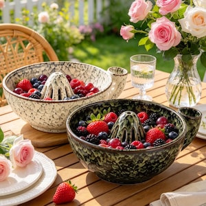 May include: Two ceramic citrus juicer bowls with handles, one in cream and one in dark green, both featuring a speckled finish and a central reamer. Each bowl is filled with fresh strawberries, blueberries, and blackberries on a wooden table.