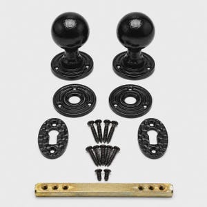 May include: A set of two black spherical door knobs with matching circular mounting plates, two oval keyhole escutcheons, ten black screws, and one brass-colored metal spindle bar. The hardware features a textured finish throughout.