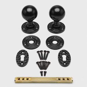 May include: A set of two black spherical door knobs with matching circular mounting plates, two oval keyhole escutcheons, a brass-colored metal spindle, and a collection of ten black screws arranged in three groups for installation hardware.
