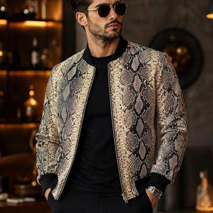 May include: A bomber jacket featuring a beige and black snakeskin pattern with a solid black ribbed collar, cuffs, and hem. The jacket is worn over a plain black t-shirt and paired with black trousers, complemented by dark sunglasses and a silver watch.