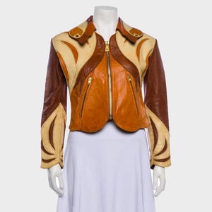 Puede incluir: Una chaqueta corta de cuero con un diseño de bloques de color en tonos crema, beige y marrón oscuro. La chaqueta cuenta con una cremallera frontal dorada, dos bolsillos con cremallera, un cuello ancho y mangas largas con paneles de cuero curvos.