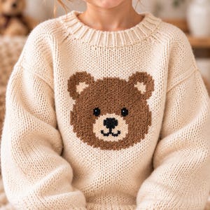Peut inclure: Un pull en tricot à manches longues de couleur crème avec un grand motif central représentant une tête d'ours marron. Le design de l'ours comprend des oreilles marron foncé, des yeux noirs et un nez noir avec une bouche détaillée.