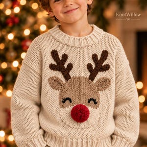 Könnte beinhalten: Ein cremefarbener, langärmeliger Strickpullover mit einem großen Rentier-Motiv auf der Vorderseite. Das Rentier hat ein dunkelbraunes Geweih, hellbraune Ohren, schwarze gestickte Augen und eine auffällige, strukturierte rote Bommelnase. Der Text KnotWillow ist sichtbar.