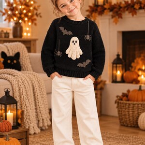May include: A black long-sleeve jumper featuring a textured cream-coloured ghost appliqué, dark grey bats, and spider motifs. The jumper is paired with off-white corduroy trousers that have a straight-leg cut and two front pockets.