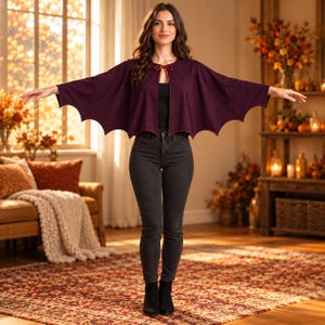 May include: A deep purple cape with a scalloped bat-wing hemline and a thin tie closure at the neckline. The garment features wide, flared sleeves that extend outward, creating a distinct silhouette. It is worn over a black top and dark jeans.