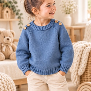 May include: A blue long-sleeved knit sweater featuring a ribbed crew neckline and cuffs, with four light brown wooden buttons positioned along the shoulder seams. The sweater has a classic stockinette stitch pattern and a relaxed fit.