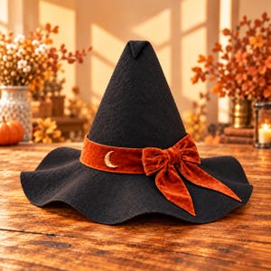 May include: A black pointed hat with a wide, wavy brim. A burnt orange velvet ribbon wraps around the base, featuring a gold-colored crescent moon charm on the left side and a large, tied velvet bow on the right side.