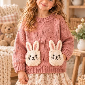 May include: A dusty rose knitted sweater featuring two cream-colored rabbit-shaped pockets on the front, paired with a cream-colored skirt adorned with a small brown floral pattern. The sweater has a ribbed crew neckline and long sleeves.